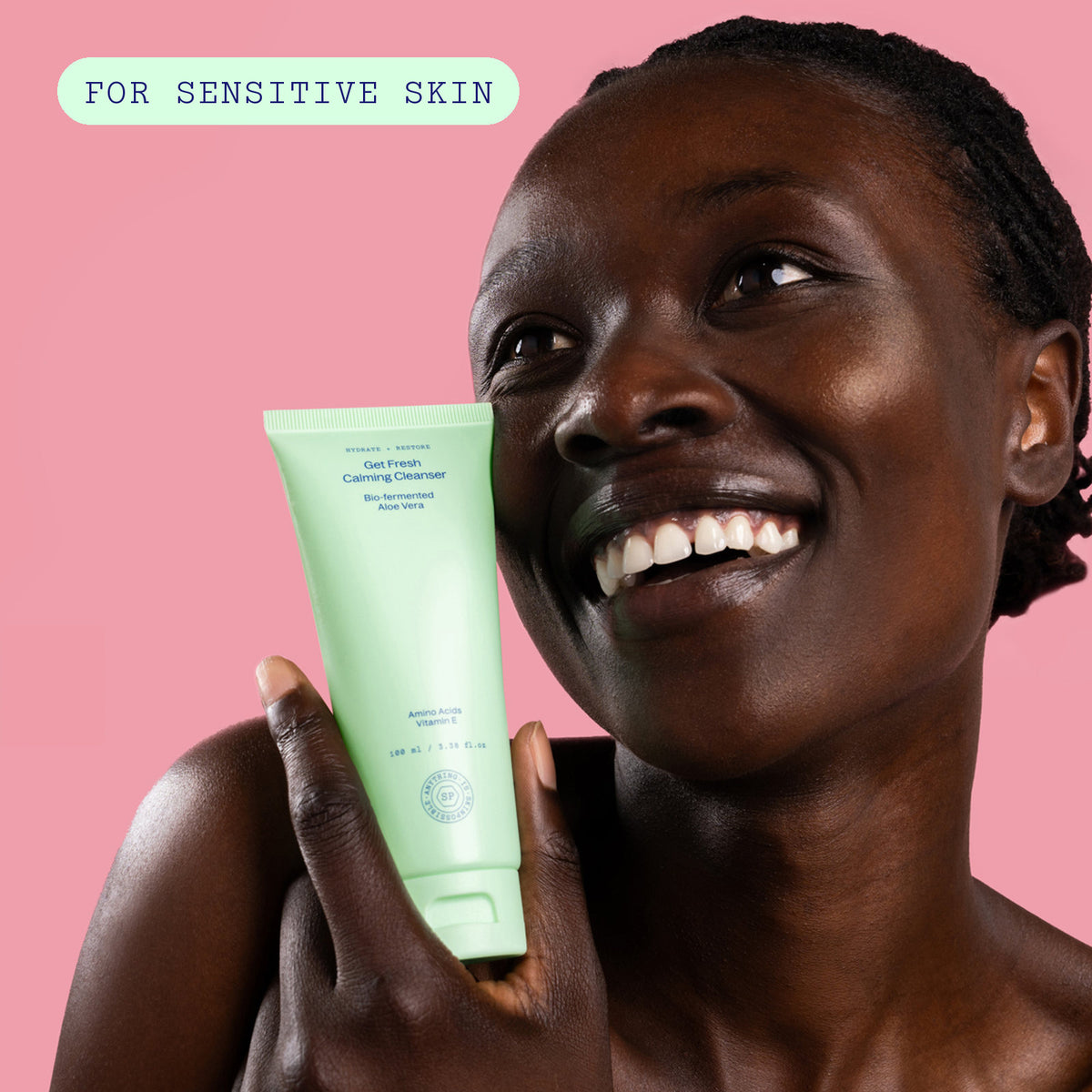 Smiling woman holding the Get Fresh Calming Cleanser to her cheek against a pink background, labelled for sensitive skin