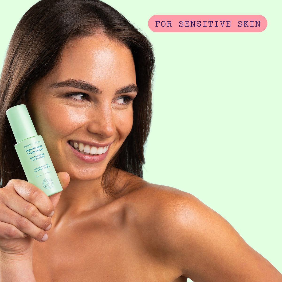 Smiling woman holding the High Achiever Super Serum bottle to her cheek against a mint green background, labelled for sensitive skin
