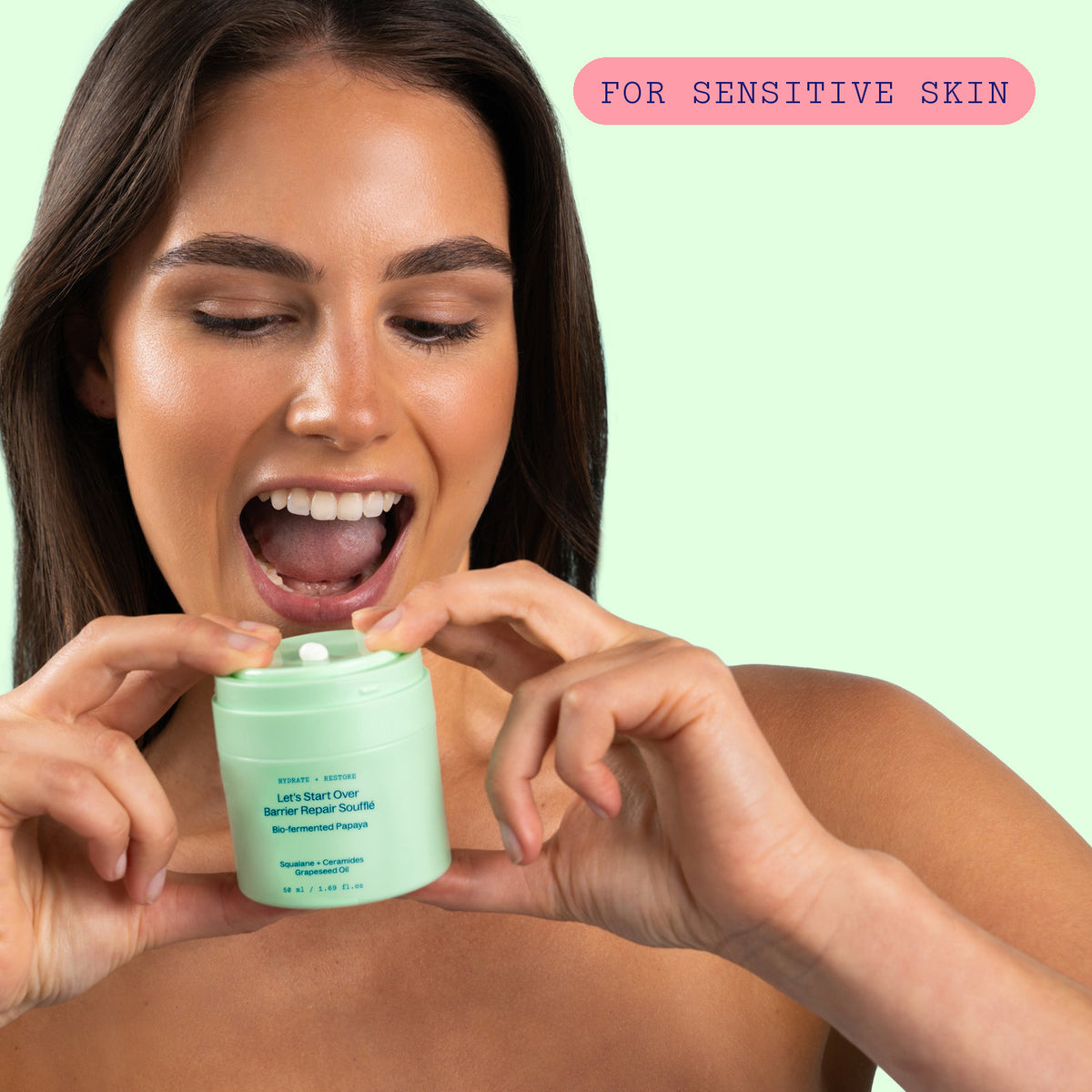 Smiling woman holding the Barrier Repair Soufflé jar open near her face against a mint green background, labelled for sensitive skin